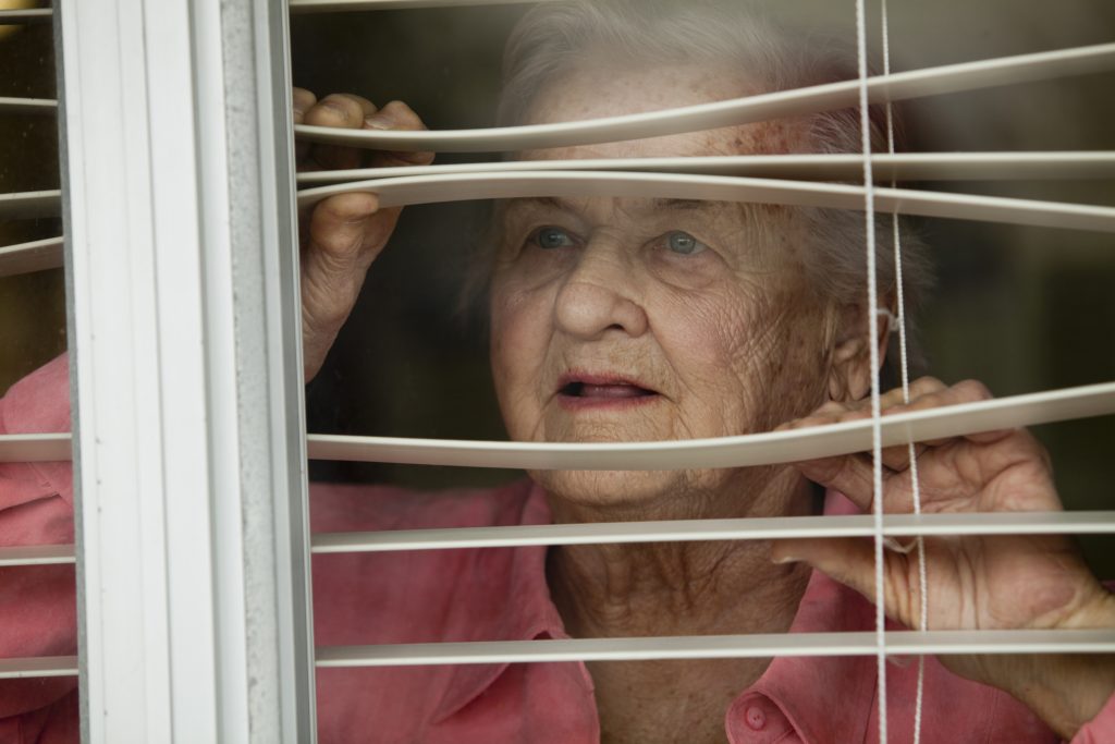 Elderly woman looking out of a window between shades.