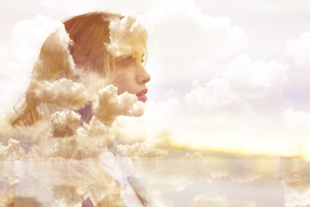 Woman with light brown hair superimposed on a dreamy landscape of a light pink sky with white clouds and a tranquil light blue body of water.