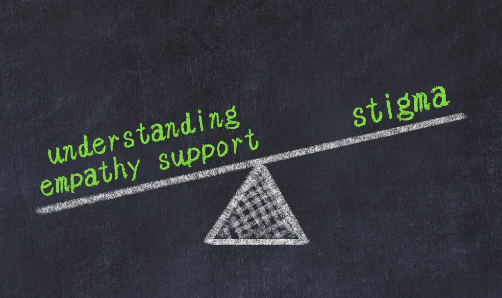 White chalk drawing of a seesaw on a blackboard texture. It's tilted to the left with three words in green chalk: 'understanding', 'empathy', and 'support'. On the right, there's a single word: 'stigma'.