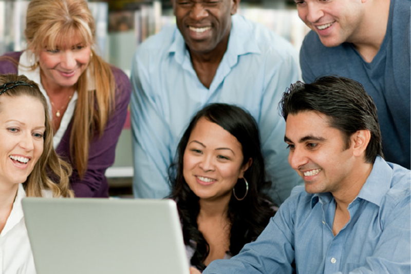 A diverse group of five smiling adults gathered around a laptop in an office setting, collaborating and sharing ideas.