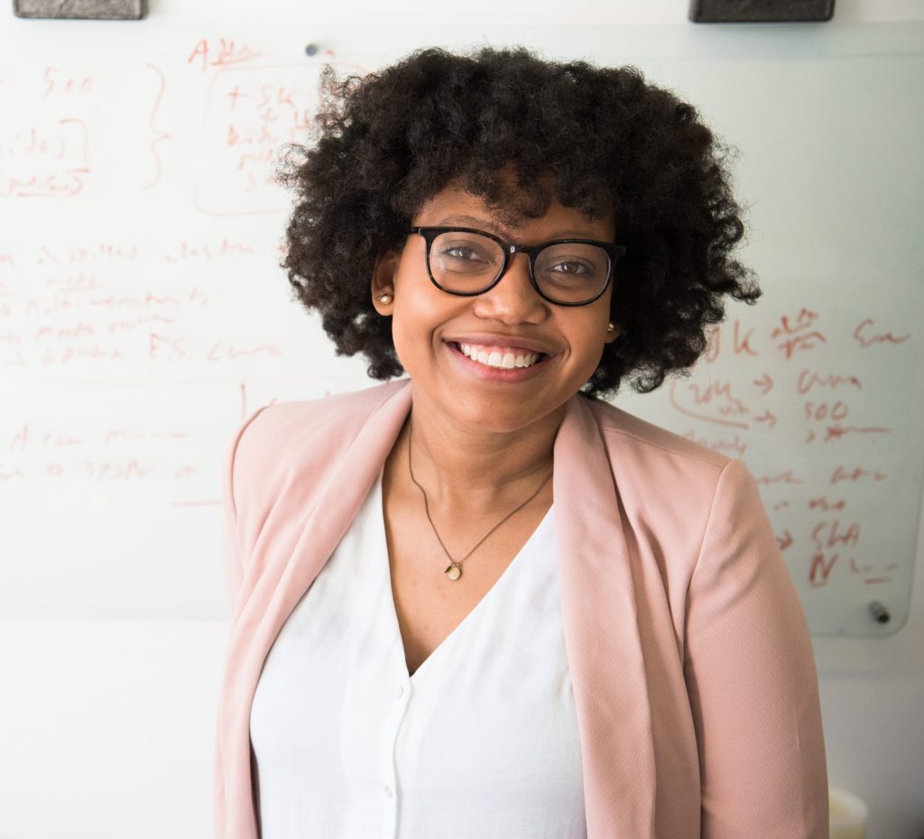 A cheerful woman with curly black hair and glasses, standing in front of a whiteboard filled with notes and diagrams.