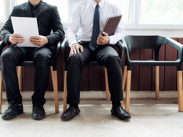 Two professionally dressed men seated in a waiting area with papers and folders in hand, waiting for a job interview.
