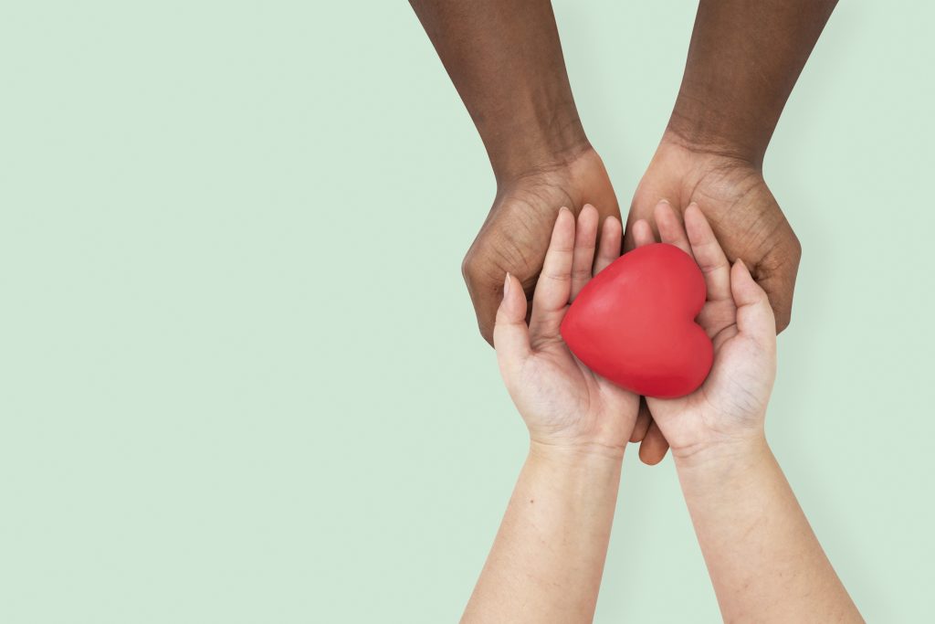 Two pairs of hands, one with a darker skin tone and one with a lighter skin tone, gently cradling a red heart-shaped object.