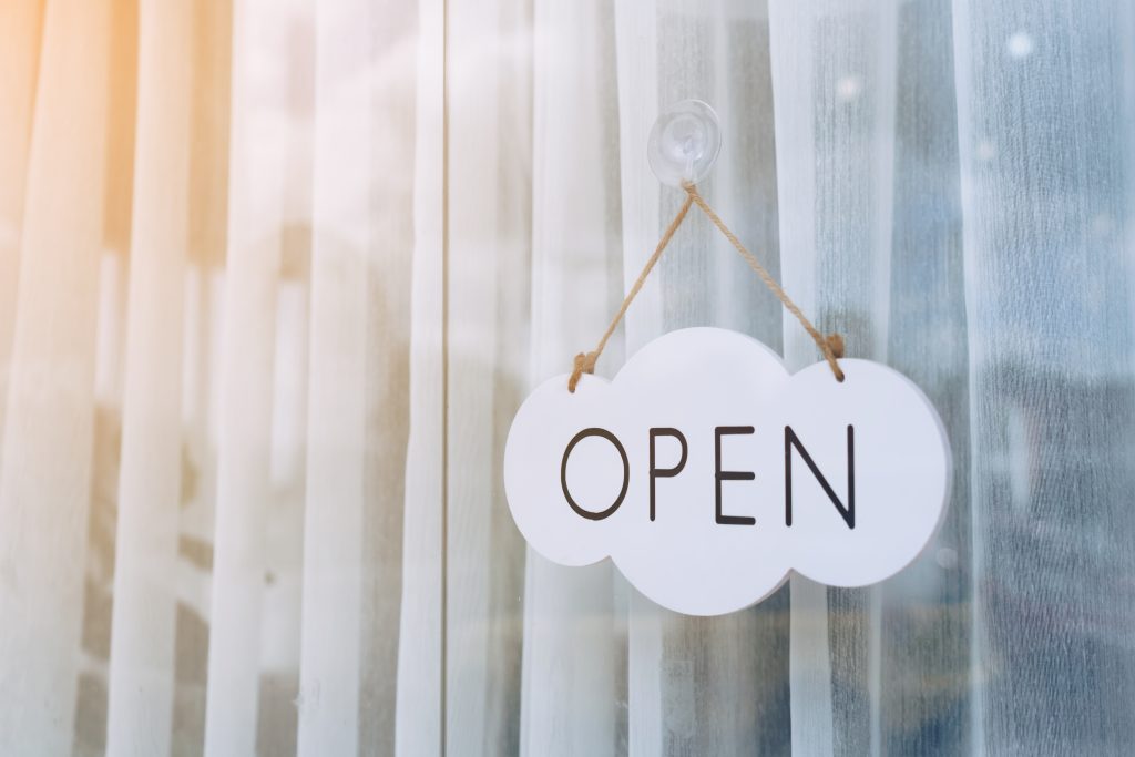 A white cloud-shaped sign with the word 'OPEN' written in black, hanging by a string on a glass door.