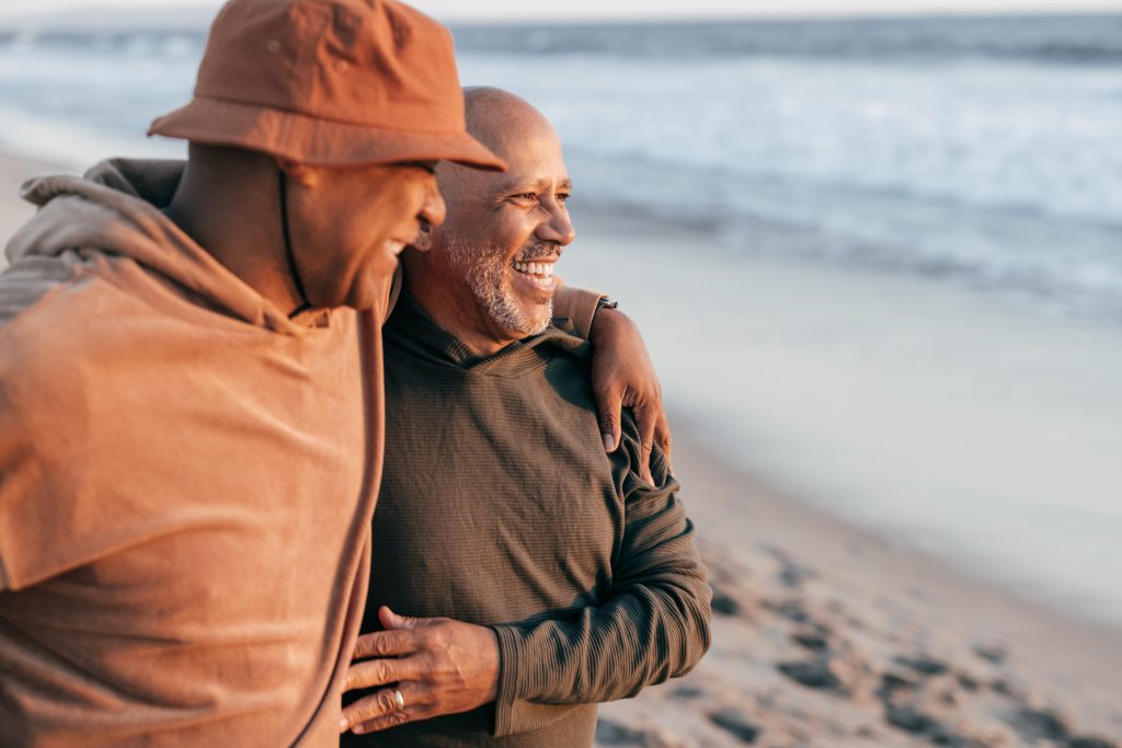 Two men on a beach looking out with arms around each other.