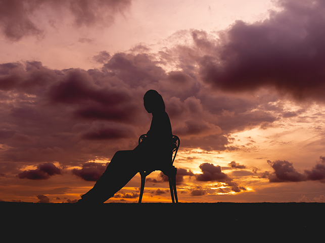 Silhouette of a solitary woman seated on a chair, set against a dramatic sunset with dark clouds and orange hues.